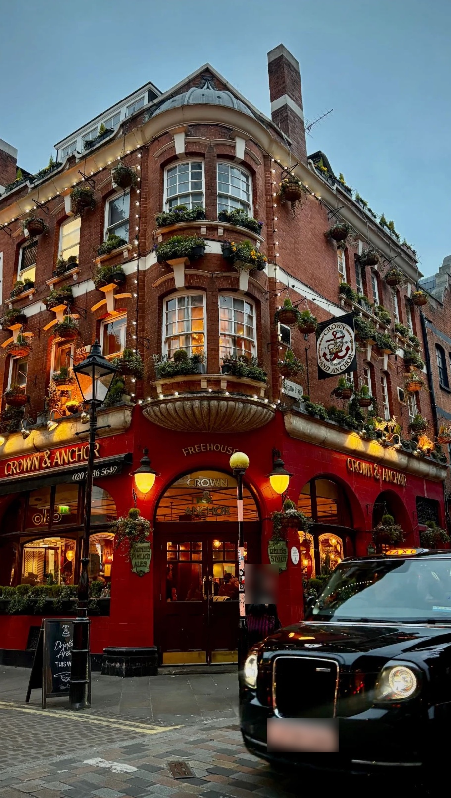 Pub londonien en briques rouges, fleuri et illuminé, avec un taxi noir devant