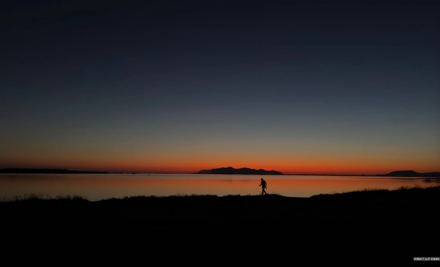 Coucher de soleil orange sur un plan d'eau, silhouette d'une personne qui marche sur la rive